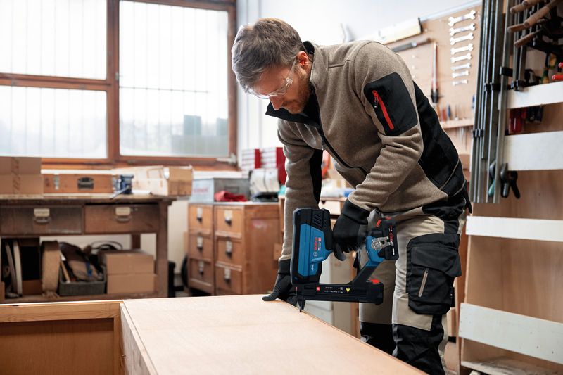 A person wearing safety equipment uses a cordless narrow crown stapler on wood.