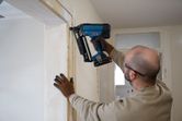 A person wearing safety equipment uses a cordless finish nailer on a door frame.