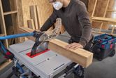 A person wearing safety equipment cuts a thick wooden beam using a table saw.