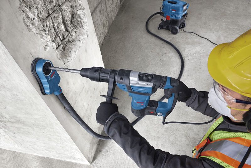 A person wearing safety equipment drills into concrete with a rotary hammer and dust extractor.
