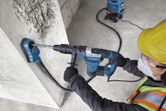 A person wearing safety equipment drills into concrete with a rotary hammer and dust extractor.