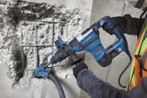 A person wearing safety equipment uses a rotary hammer to drill into concrete.