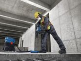 A worker wearing safety equipment uses a rotary hammer drill on a concrete floor.