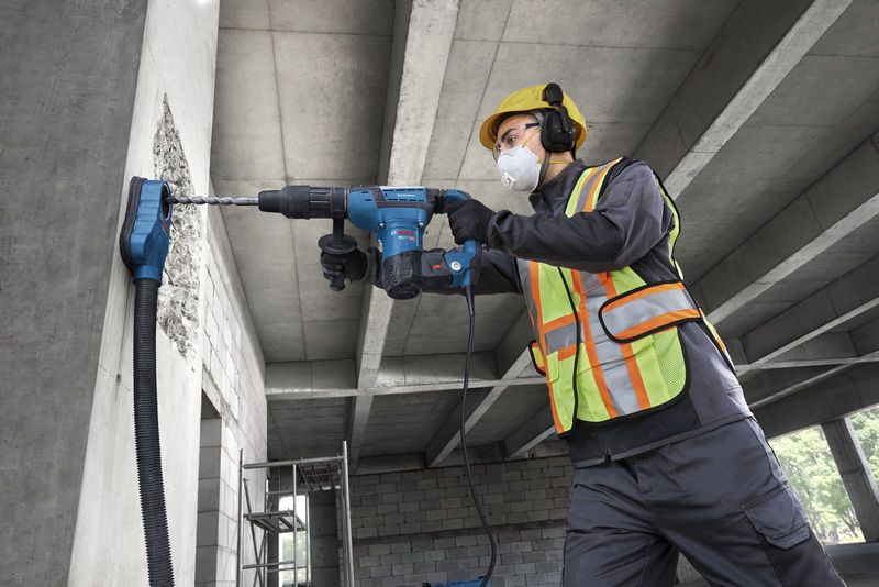 A worker wearing safety equipment drills into a concrete wall with a rotary hammer.