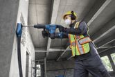 A worker wearing safety equipment drills into a concrete wall with a rotary hammer.
