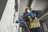 A worker wearing safety equipment uses a rotary hammer to drill into a concrete wall.