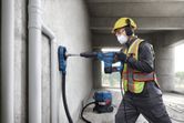 A person wearing safety equipment uses a rotary hammer to drill into a concrete wall.