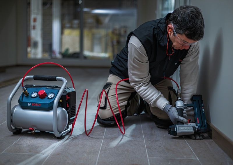 A person wearing safety equipment uses a pneumatic tool to fasten trim along a wall.