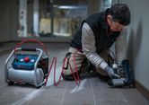 A person wearing safety equipment uses a pneumatic tool to fasten trim along a wall.