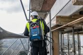 Person wearing safety equipment stands on scaffolding outdoors with a blue backpack vacuum system.