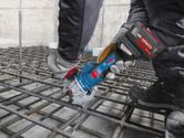 A person wearing safety equipment uses a cordless rebar cutter on a steel grid.
