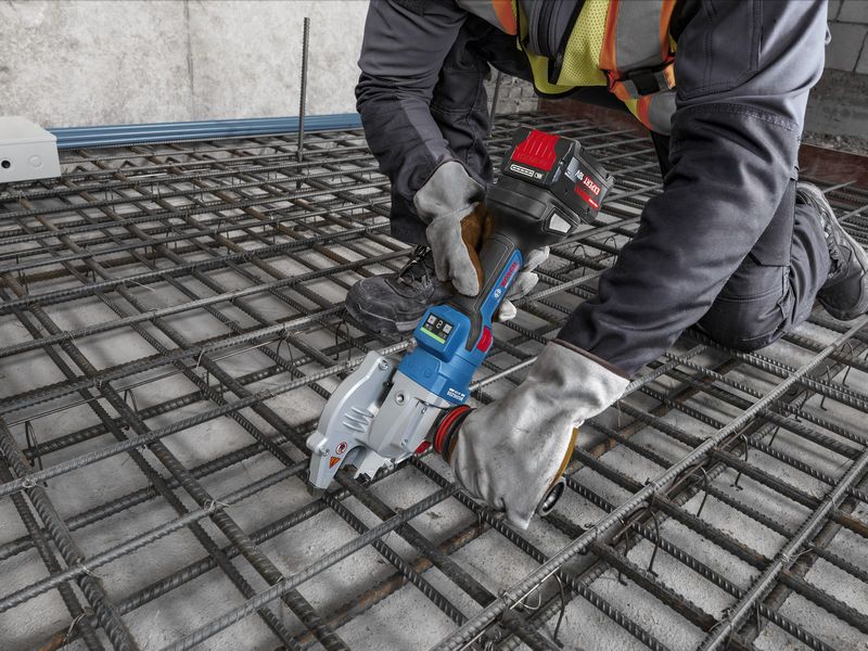 A person wearing safety equipment uses a cordless tool to cut rebar on a construction site.