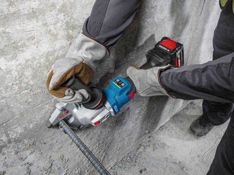 A person wearing safety equipment uses a cordless tool to cut rebar next to a concrete wall.
