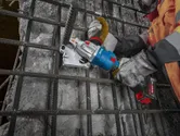 A person wearing safety equipment uses a cordless tool to cut through rebar on a concrete surface.