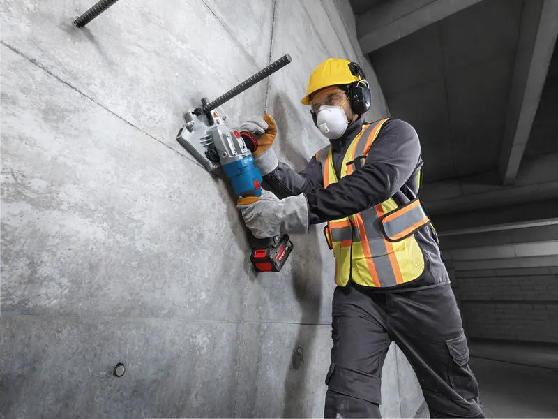 A person wearing safety equipment uses a cordless rebar cutter on a concrete wall.