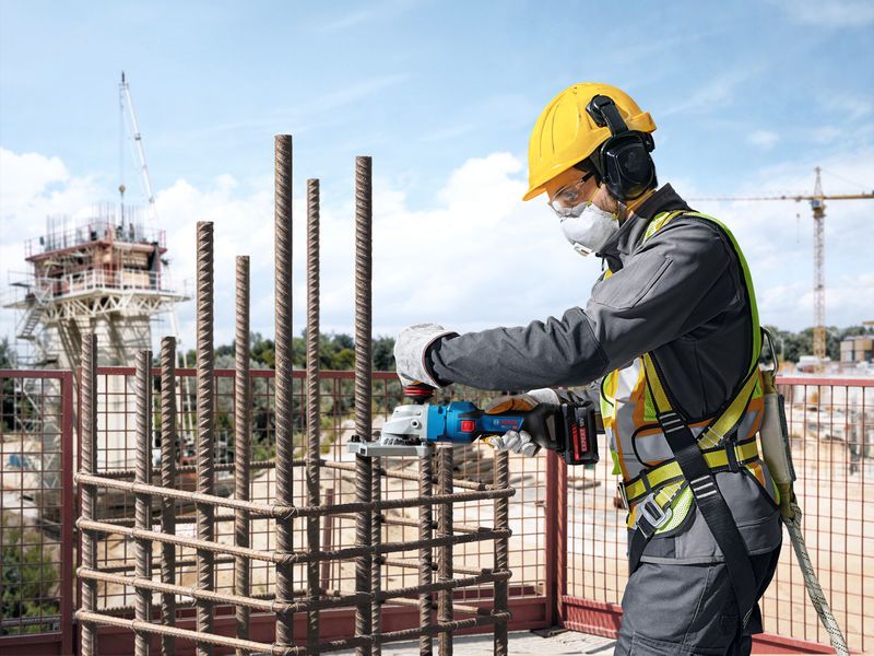 A person wearing safety equipment cuts rebar at a construction site with a cordless tool.