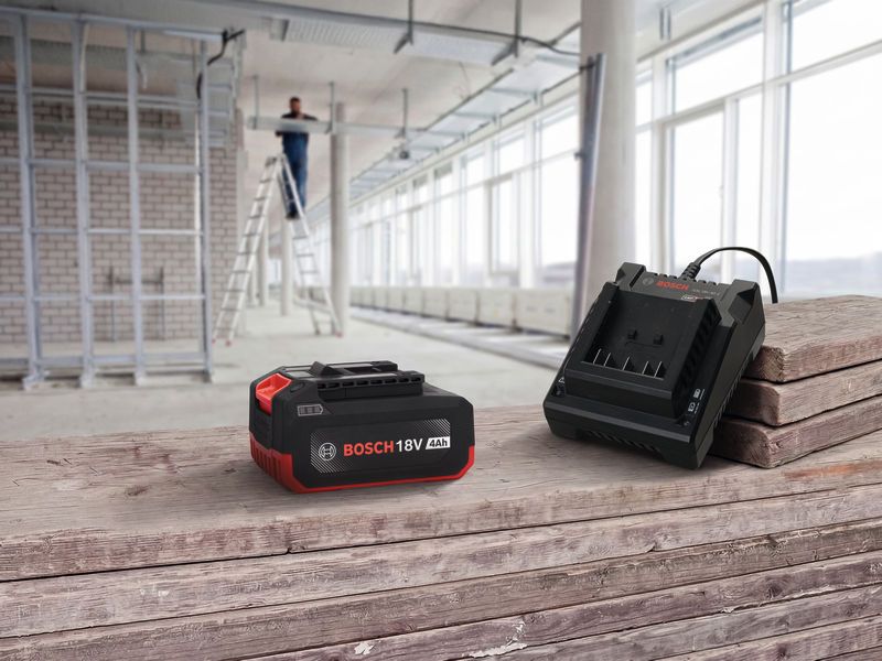 Rechargeable battery and charger on a wooden surface, with a person working on a ladder in the background.