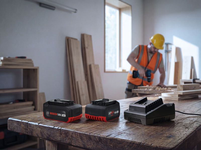 A person wearing safety equipment drills wood in a workshop with power tool batteries on a table.