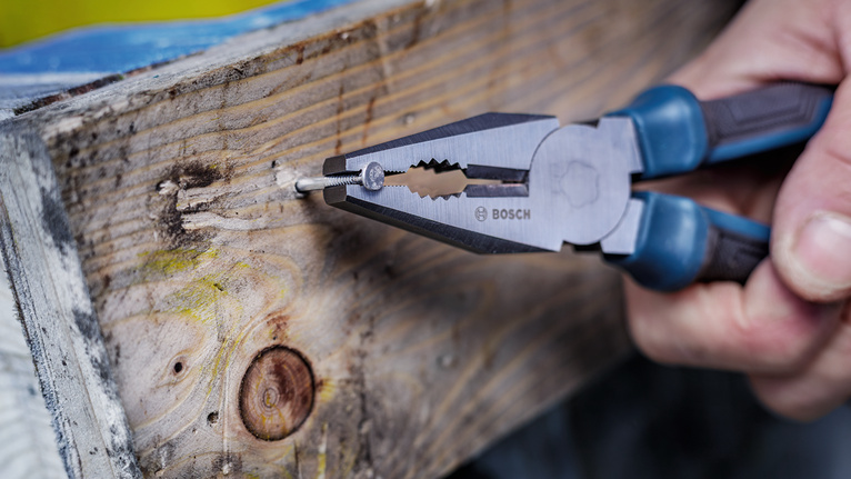 Person uses pliers to grip a nail in a wooden board.