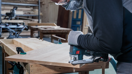 Person wearing safety equipment cuts wood with a jigsaw in a workshop.
