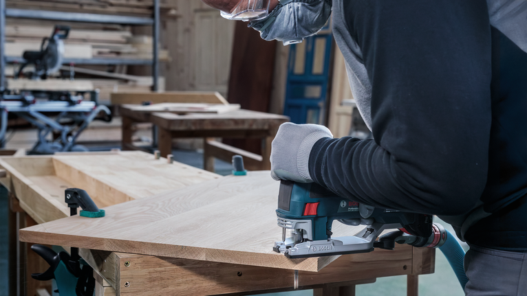 Person wearing safety equipment cuts wood with a jigsaw in a workshop.