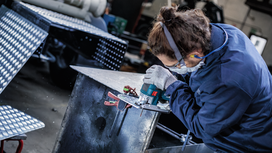 Person wearing safety equipment grinds metal using a power tool.