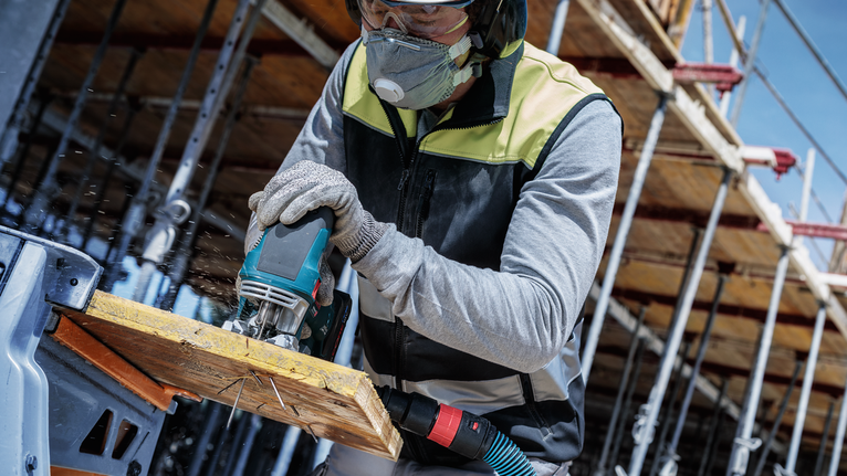 A person wearing safety equipment cuts wood with a power saw at a construction site.