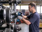 A person wearing safety equipment uses an impact wrench to tighten bolts on machinery.