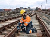 A worker wearing safety equipment uses an impact wrench to fasten bolts on railway tracks.