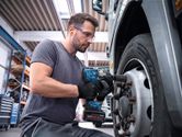 A person wearing safety equipment uses an impact wrench to tighten lug nuts on a truck wheel.