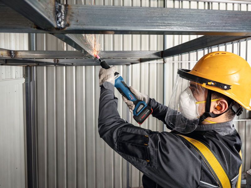 A person wearing safety equipment uses a cordless straight grinder to grind a metal beam.