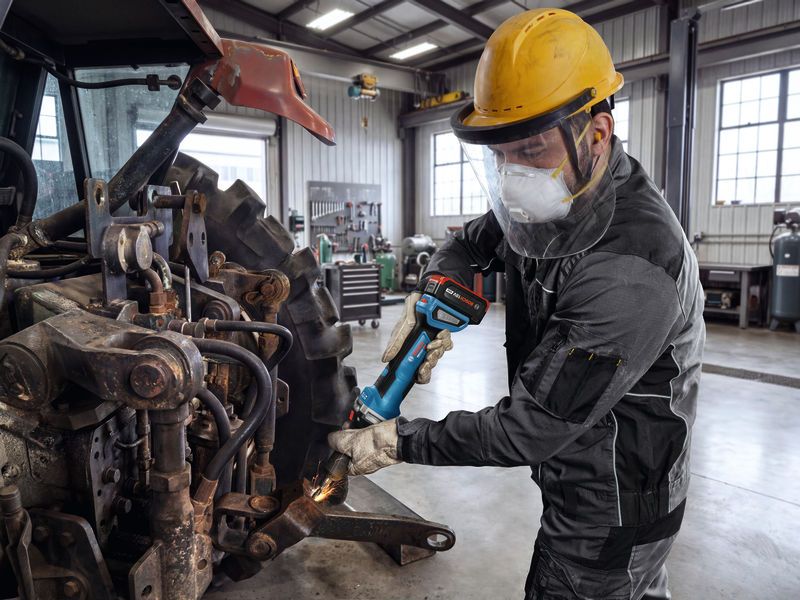 A person wearing safety equipment grinds metal on farm machinery in a workshop.