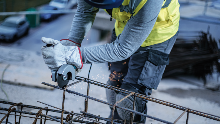 A person wearing safety equipment uses a grinder to cut steel rebar.