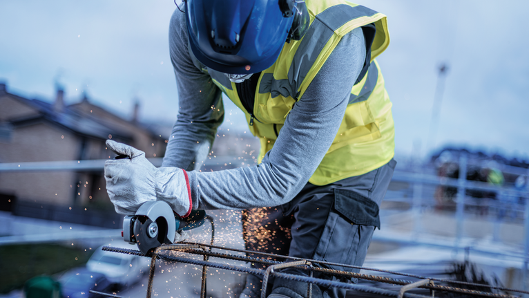 Person wearing safety equipment uses an angle grinder to cut metal rebar, producing sparks.