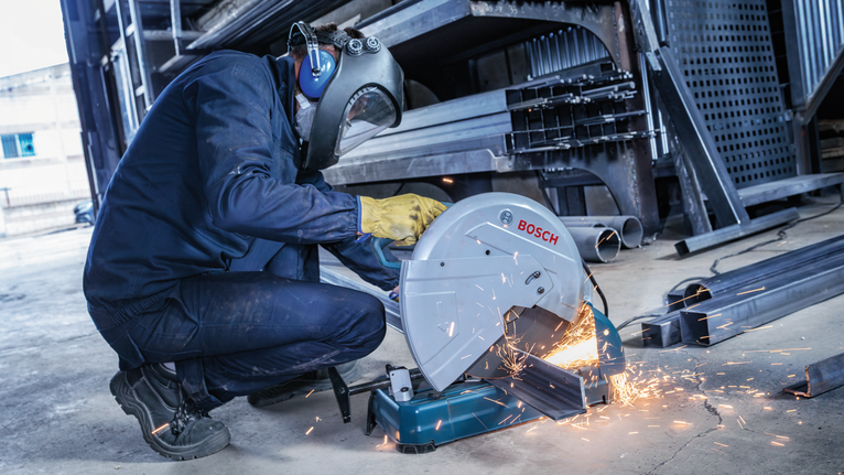 A person wearing safety equipment cuts metal beams, creating sparks on a workshop floor.