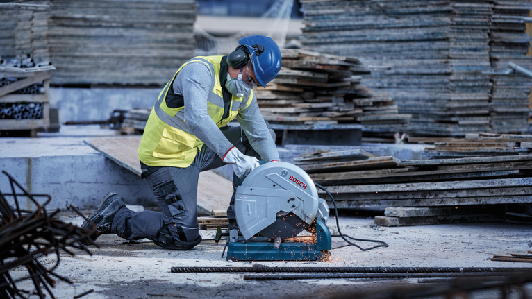 Person wearing safety equipment cuts metal bars with a heavy-duty saw at a construction site.