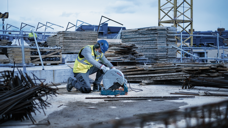A person wearing safety equipment cuts metal rods with a circular saw at a construction site.