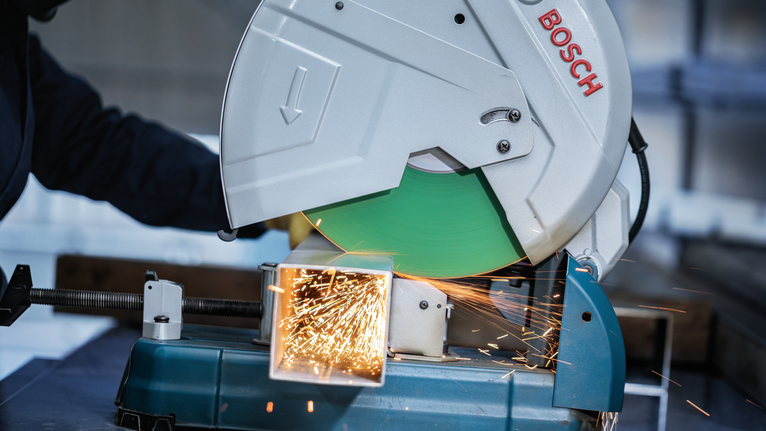Person wearing safety equipment cuts a metal beam with a chop saw, sparks flying.