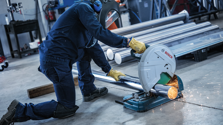 A person wearing safety equipment cuts a metal pipe with a circular saw, creating sparks.