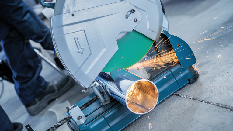 A person wearing safety equipment uses a metal chop saw to cut a pipe, creating sparks.