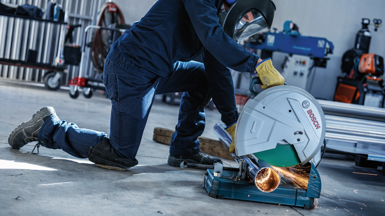 A person wearing safety equipment cuts a metal pipe with a powered chop saw.