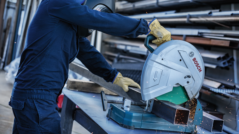 A person wearing safety equipment cuts a metal beam with a chop saw.
