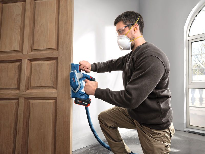 A person wearing safety equipment uses a cordless planer on a wooden door.