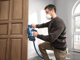 A person wearing safety equipment uses a cordless planer on a wooden door.