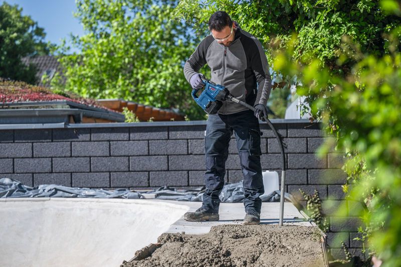 A person wearing safety equipment uses a power tool to compact concrete outdoors.