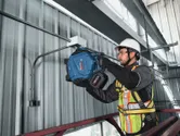 A person wearing safety equipment drills into a metal wall inside an industrial building.