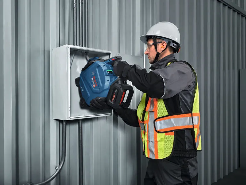 A person wearing safety equipment drills into an electrical box mounted on a metal wall.