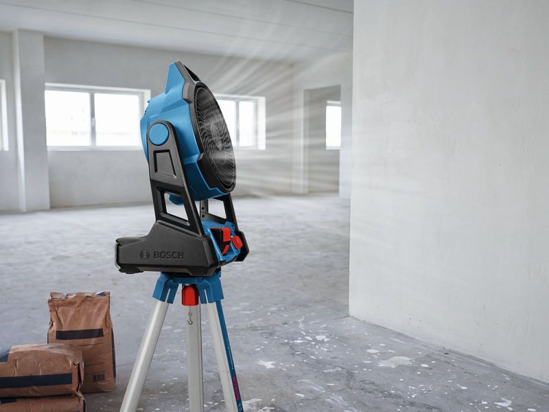 Portable jobsite fan on a tripod blows air in an empty room with construction bags.