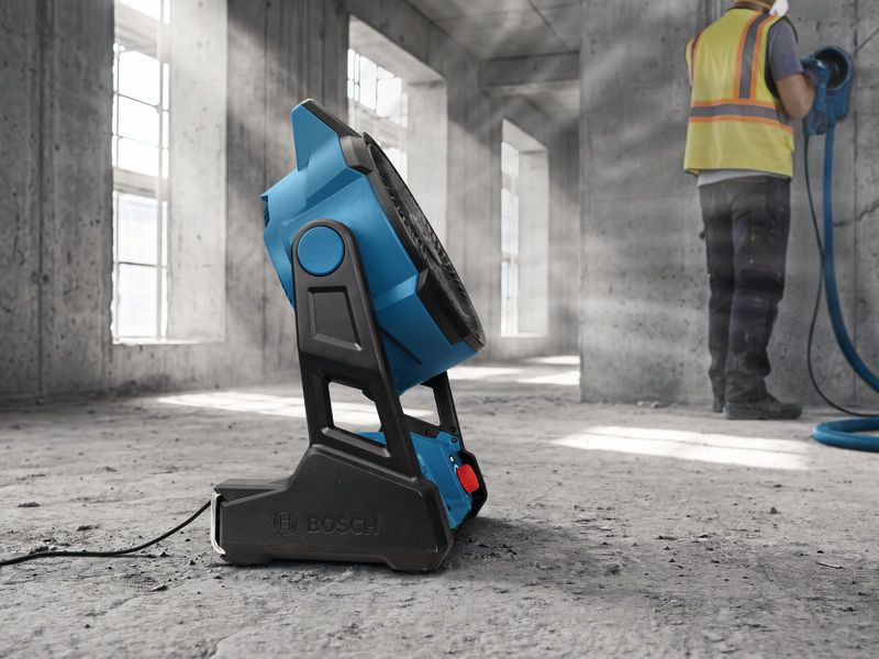 Portable jobsite fan on a concrete floor, person wearing safety equipment uses a grinder nearby.