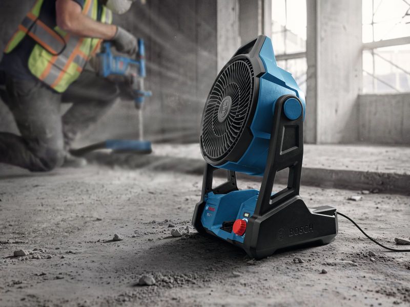 A person wearing safety equipment operates a power drill beside a portable jobsite fan.